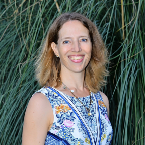 Photo of Julia from the shoulders up, standing in front of long green grass. She has shoulder length dark blonde hair and wears a blue paisley pattered vest top.