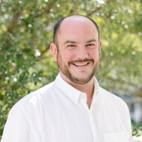 Photo of Mark standing in front of a green background with trees, smiling. He wears a white shirt.